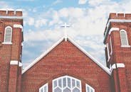 Red brick church with a cross and arched windows under a blue sky with clouds.